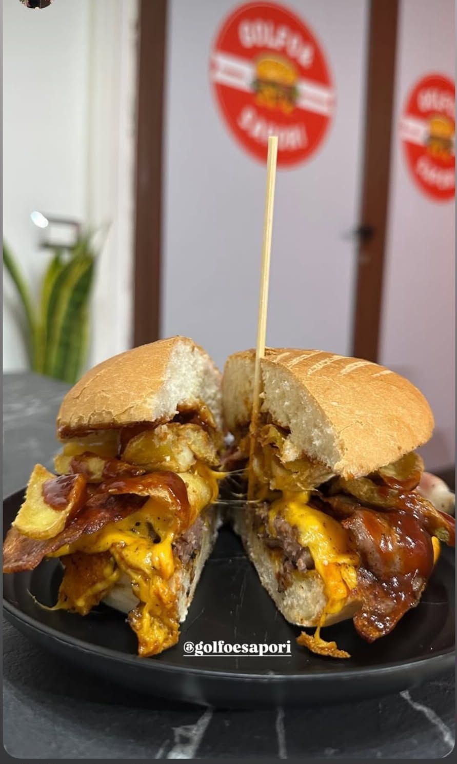 Due metà di cheeseburger impilate con formaggio fuso e pancetta su un piatto nero, con una bandierina fatta con uno stuzzicadenti sullo sfondo.