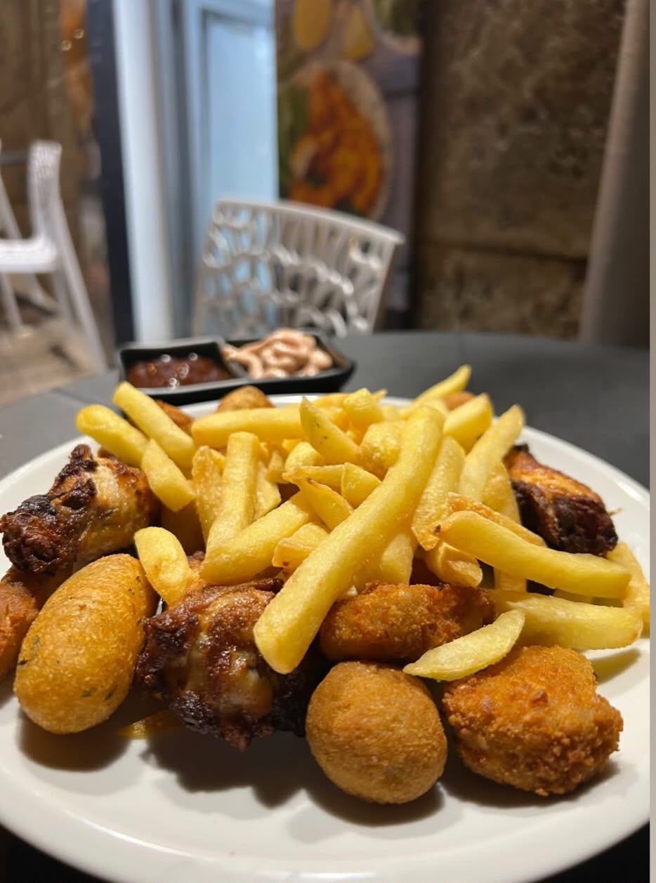 Un vassoio di patatine fritte e antipasti impanati su un tavolo da patio, con carne alla griglia sullo sfondo.