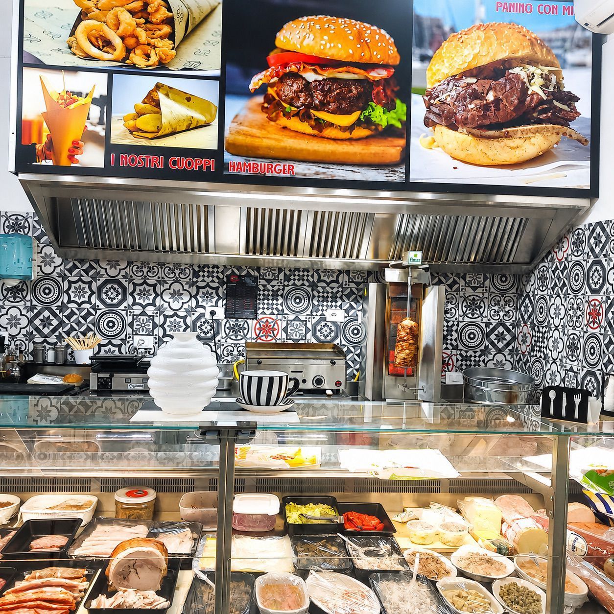 Banco gastronomia con pasticcini e dessert esposti sotto lavagne con menù, e hamburger e fritti in bella vista in una vetrina.