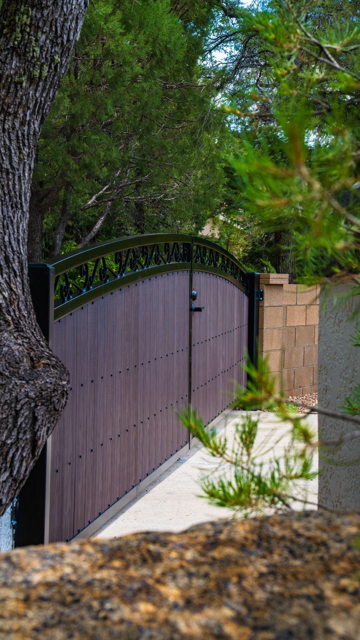 Wooden gate with decorative top, viewed through foliage, next to a stone wall.