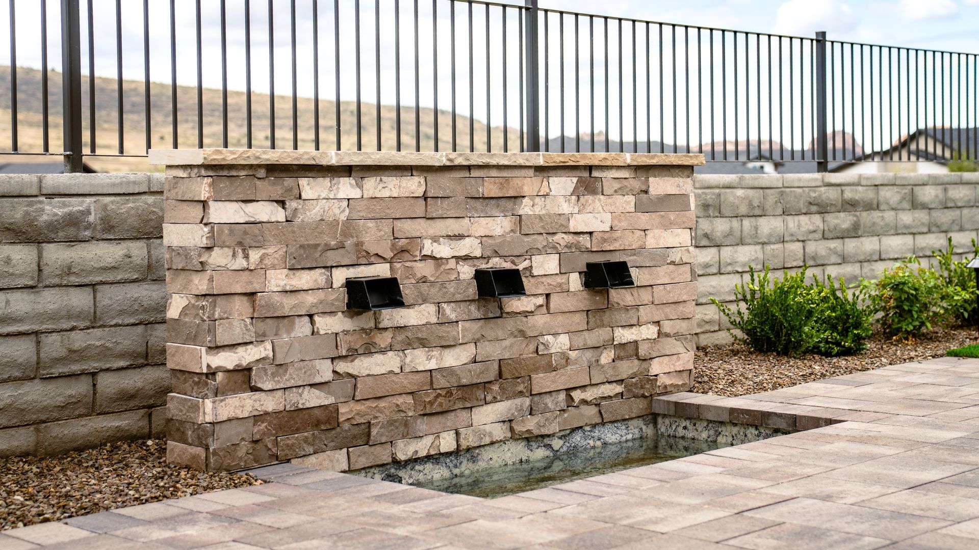 Brick water fountain with three spouts, surrounded by stone and a black metal fence.