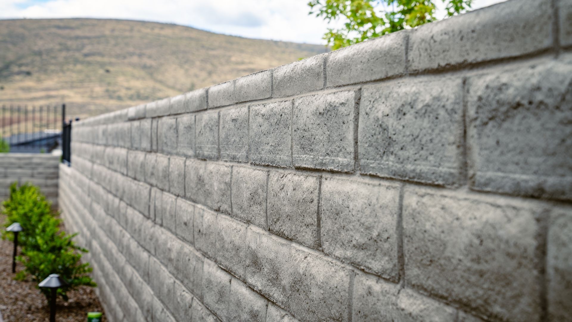 Gray concrete retaining wall in a yard, with a hillside and plants in the background.
