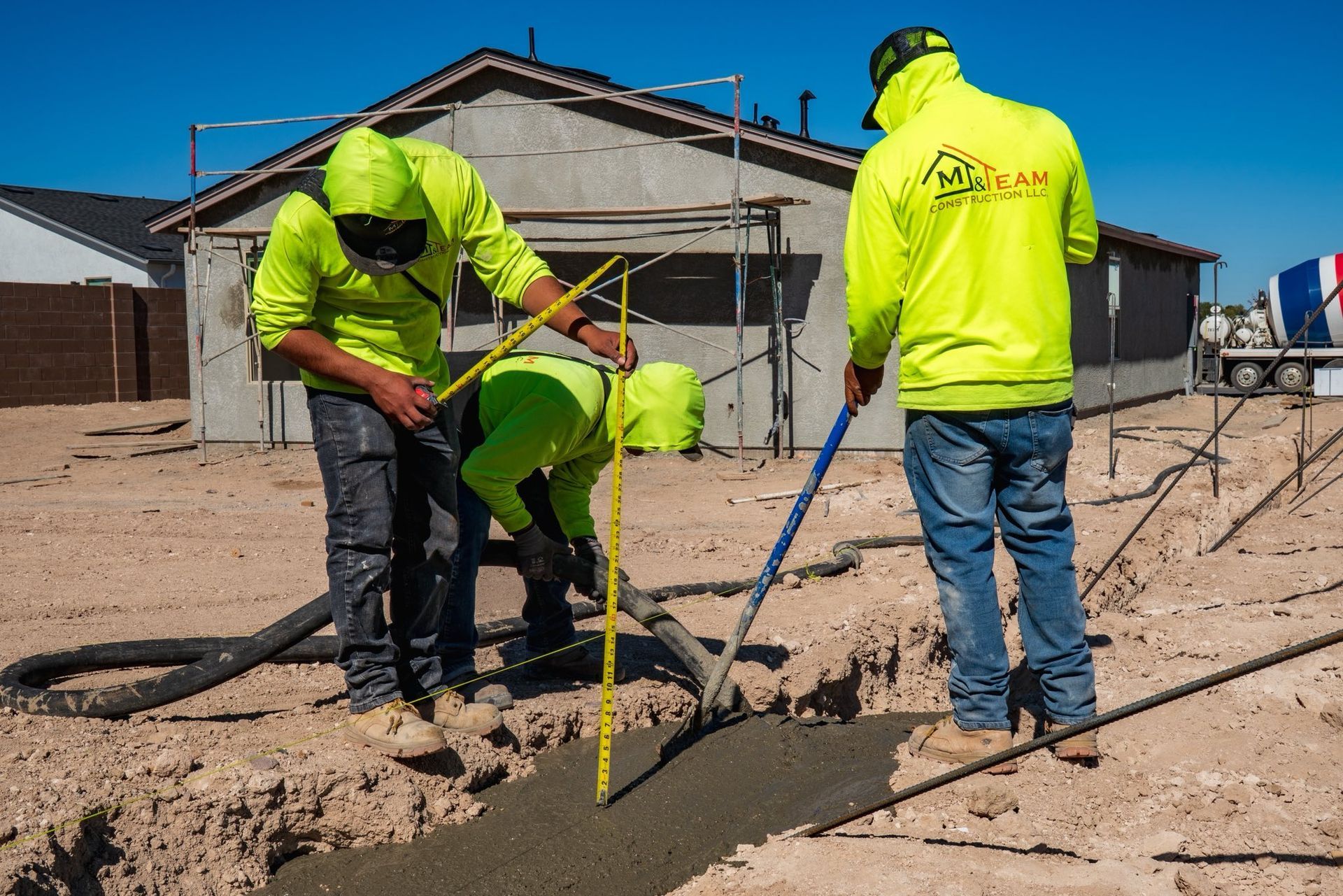 Construction workers pouring concrete into a trench at a building site. Workers wear neon jackets in a sunny environment.