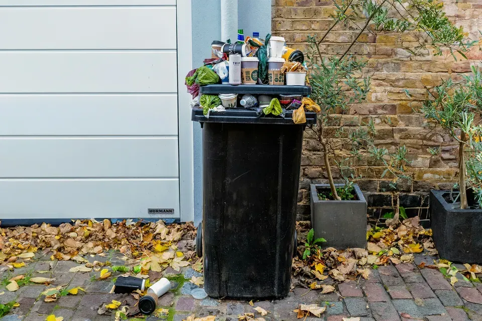 A black garbage bin with lots of trash sitting in front of a garage door