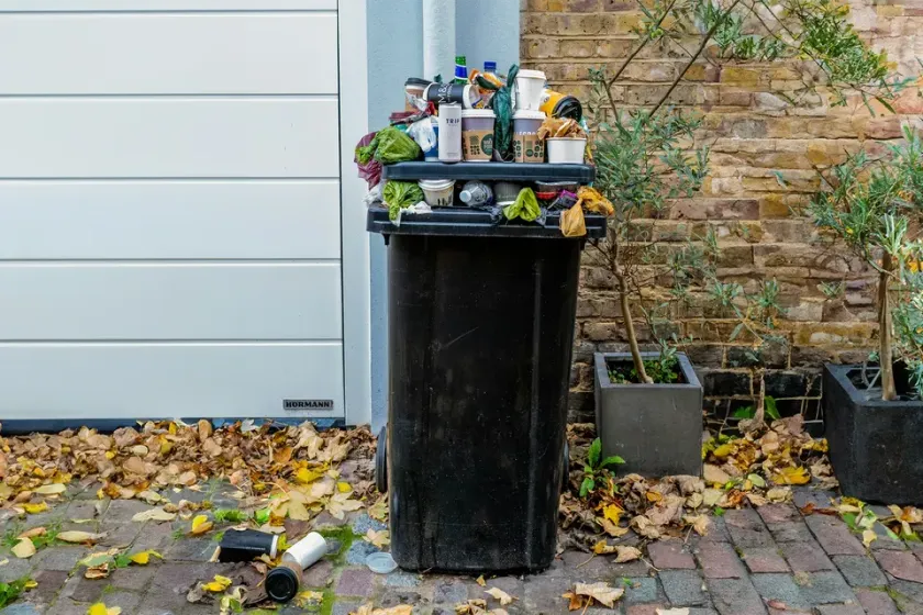 A black trash can filled with garbage is sitting on a brick sidewalk in front of a garage door in Vineland, NJ
