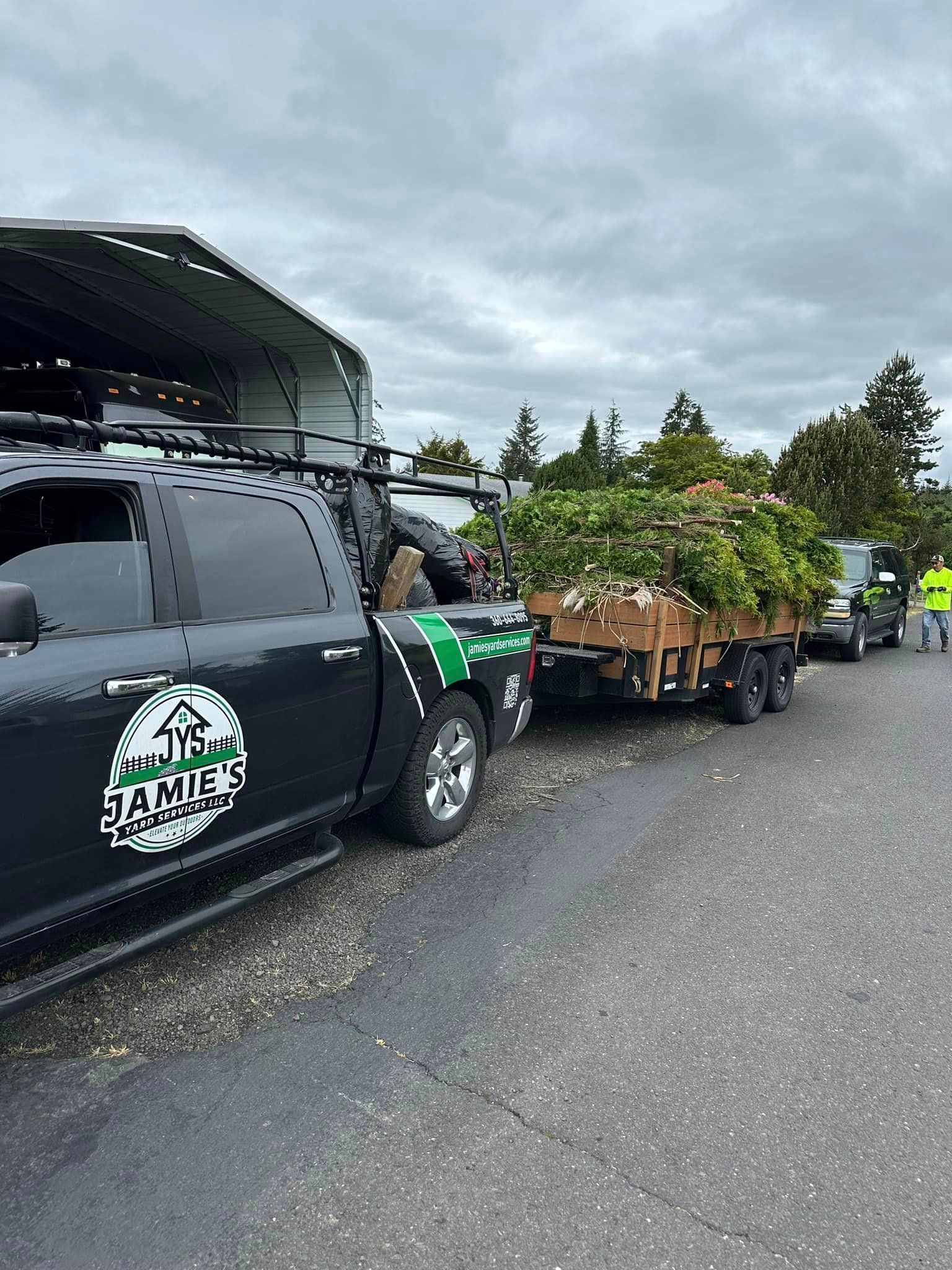 Pickup truck towing a trailer of potted plants on a gravel lot under cloudy skies