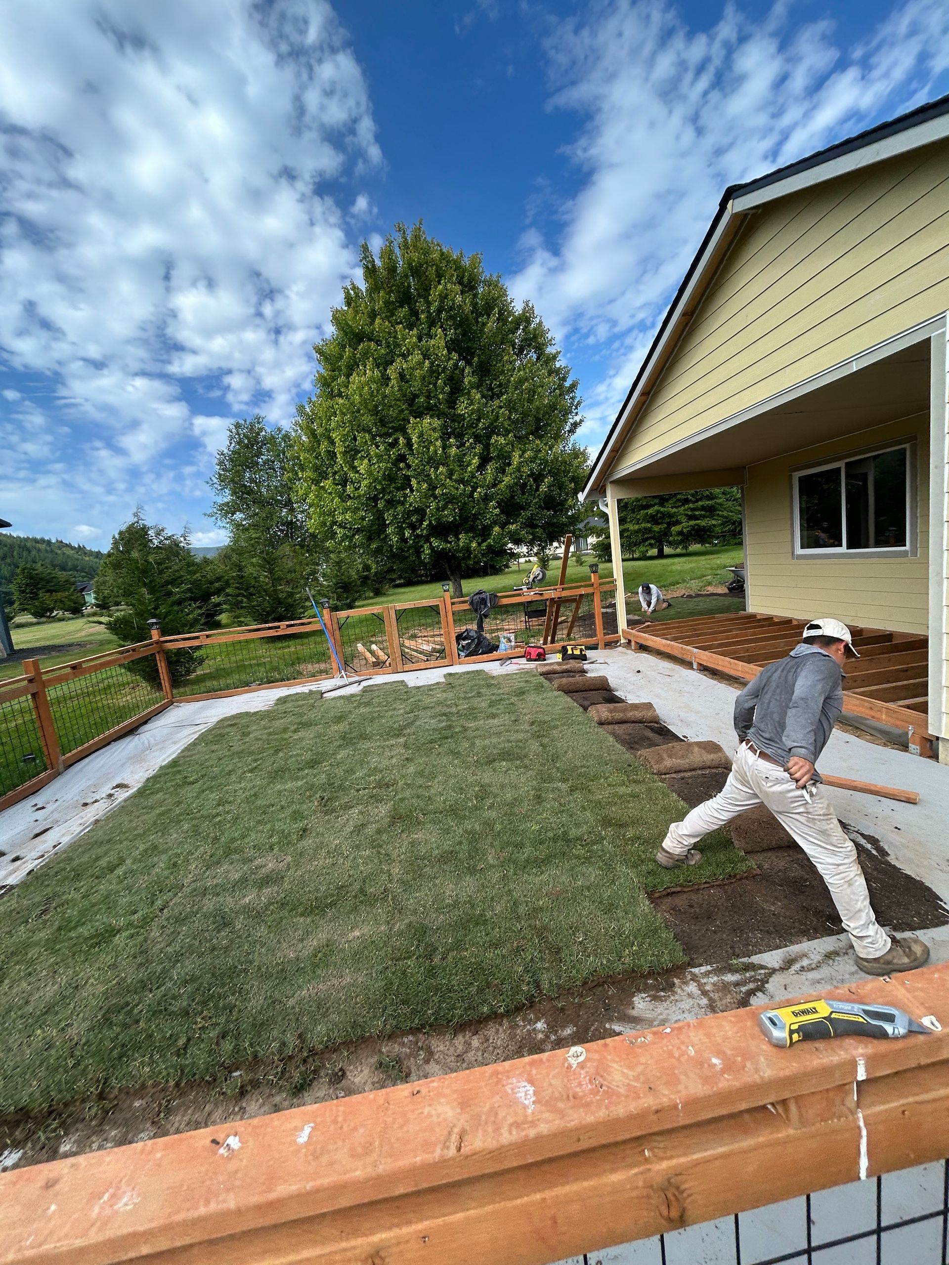Worker installing sod in a backyard beside a beige house and wooden deck under a blue sky.