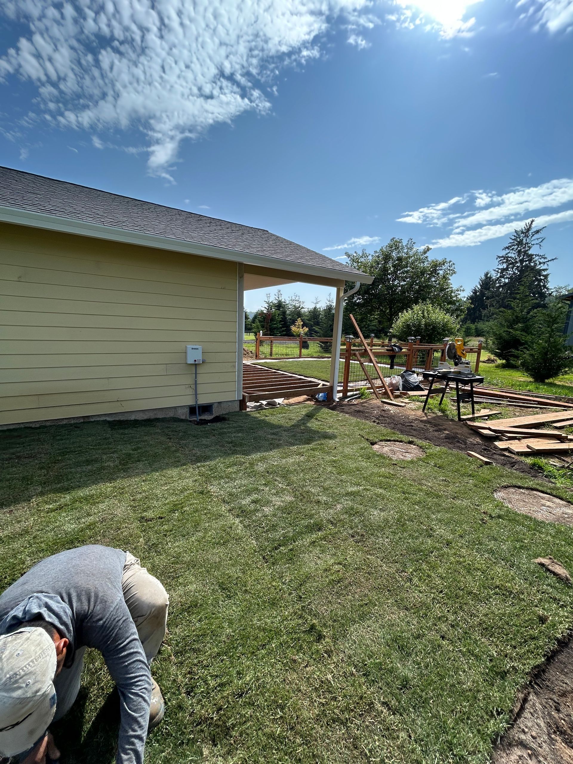 Backyard with a house, patio, garden beds, and a person working in the grass under a blue sky