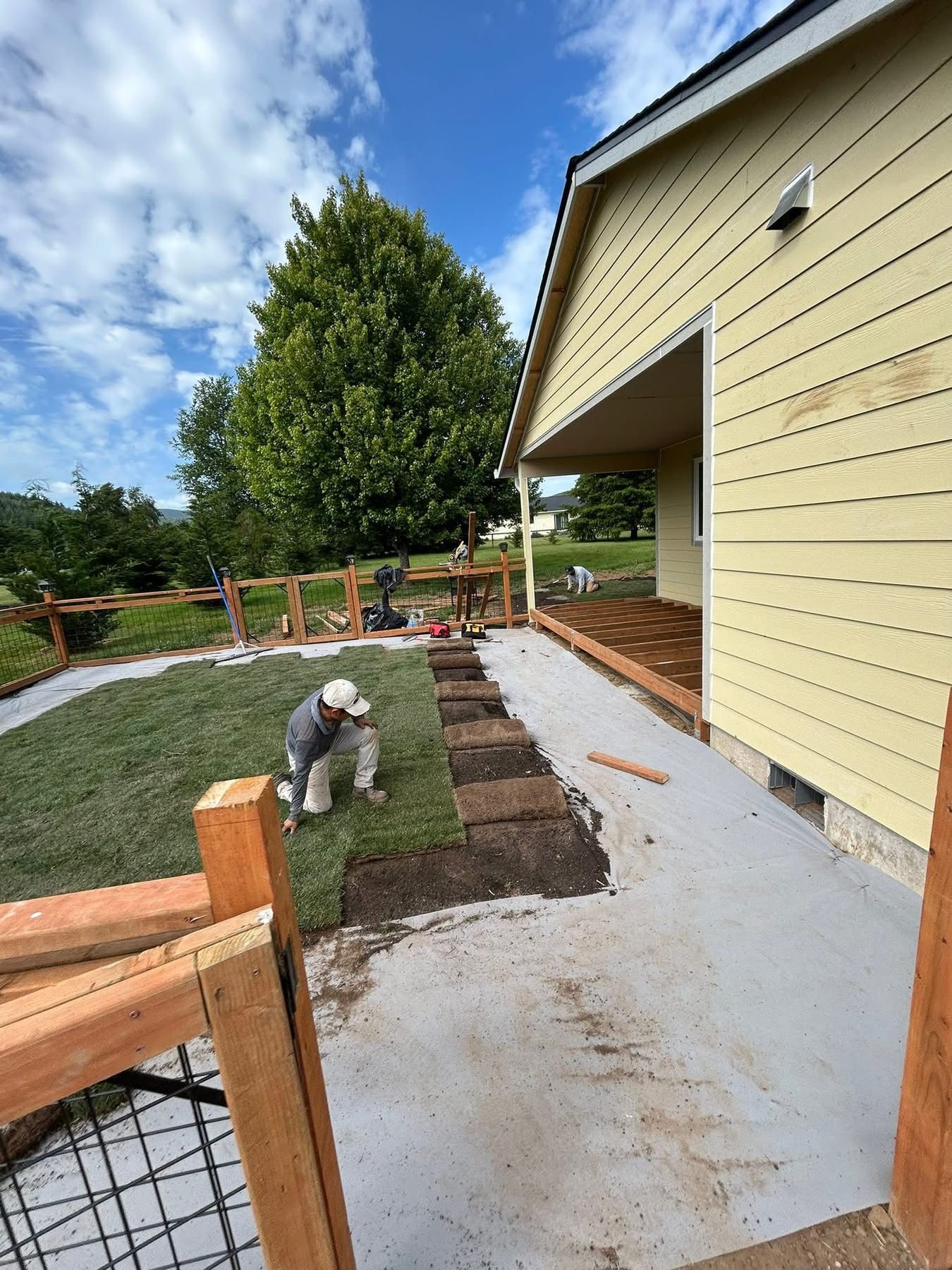 Person digging a garden bed beside a yellow house with a concrete patio and wooden fence