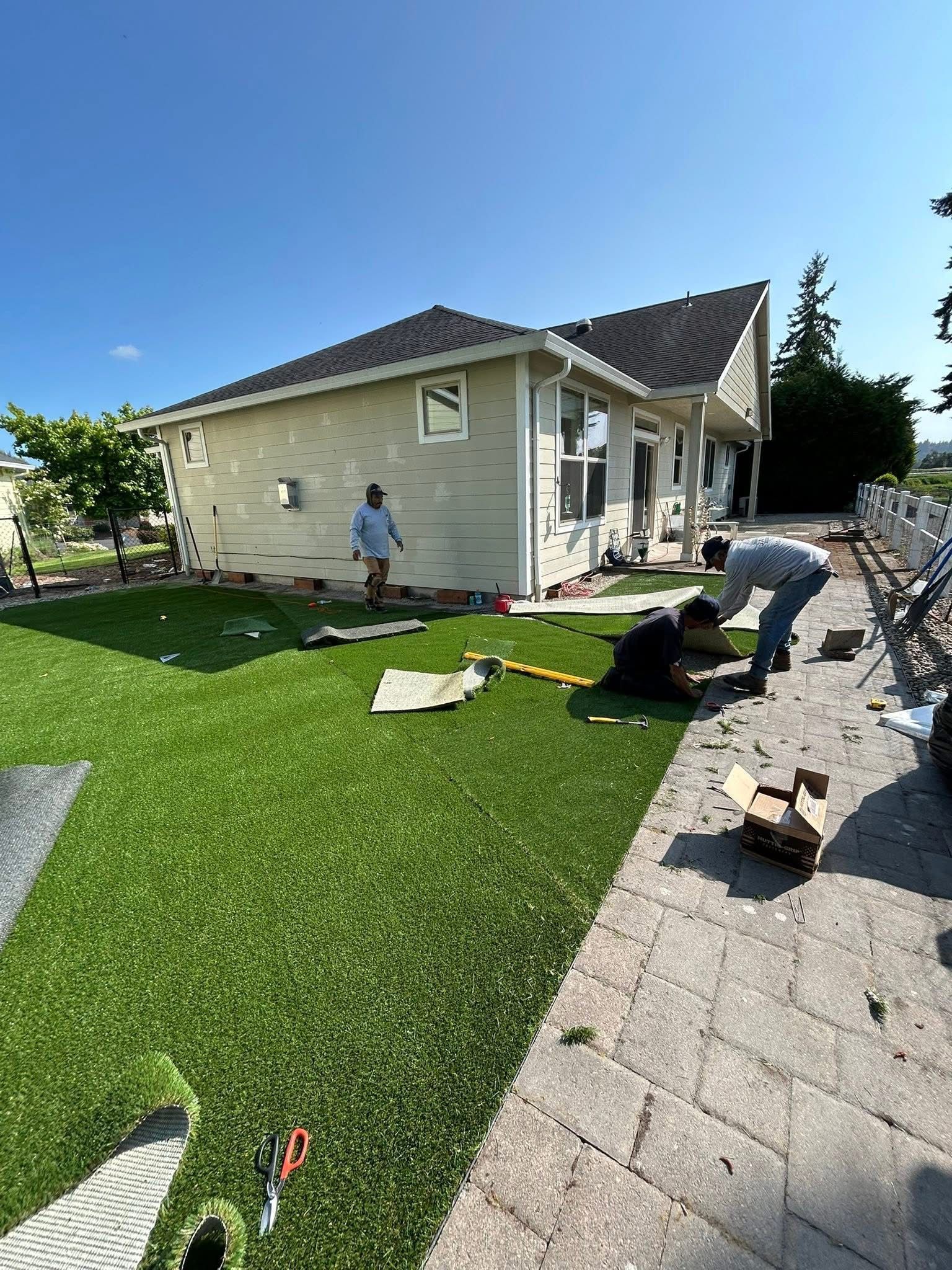 Workers installing artificial turf beside a beige house under a clear blue sky