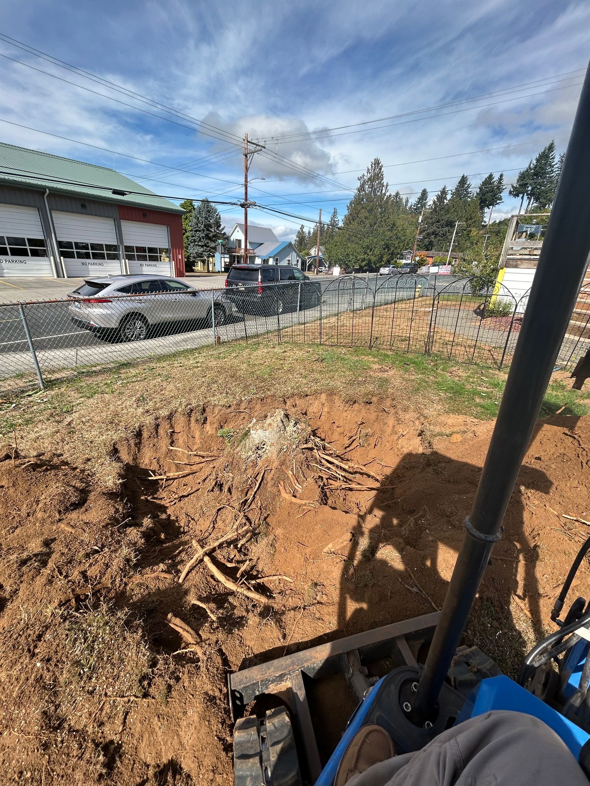 Dug-up dirt lot beside a road, with parked cars, trees, and a blue sky overhead