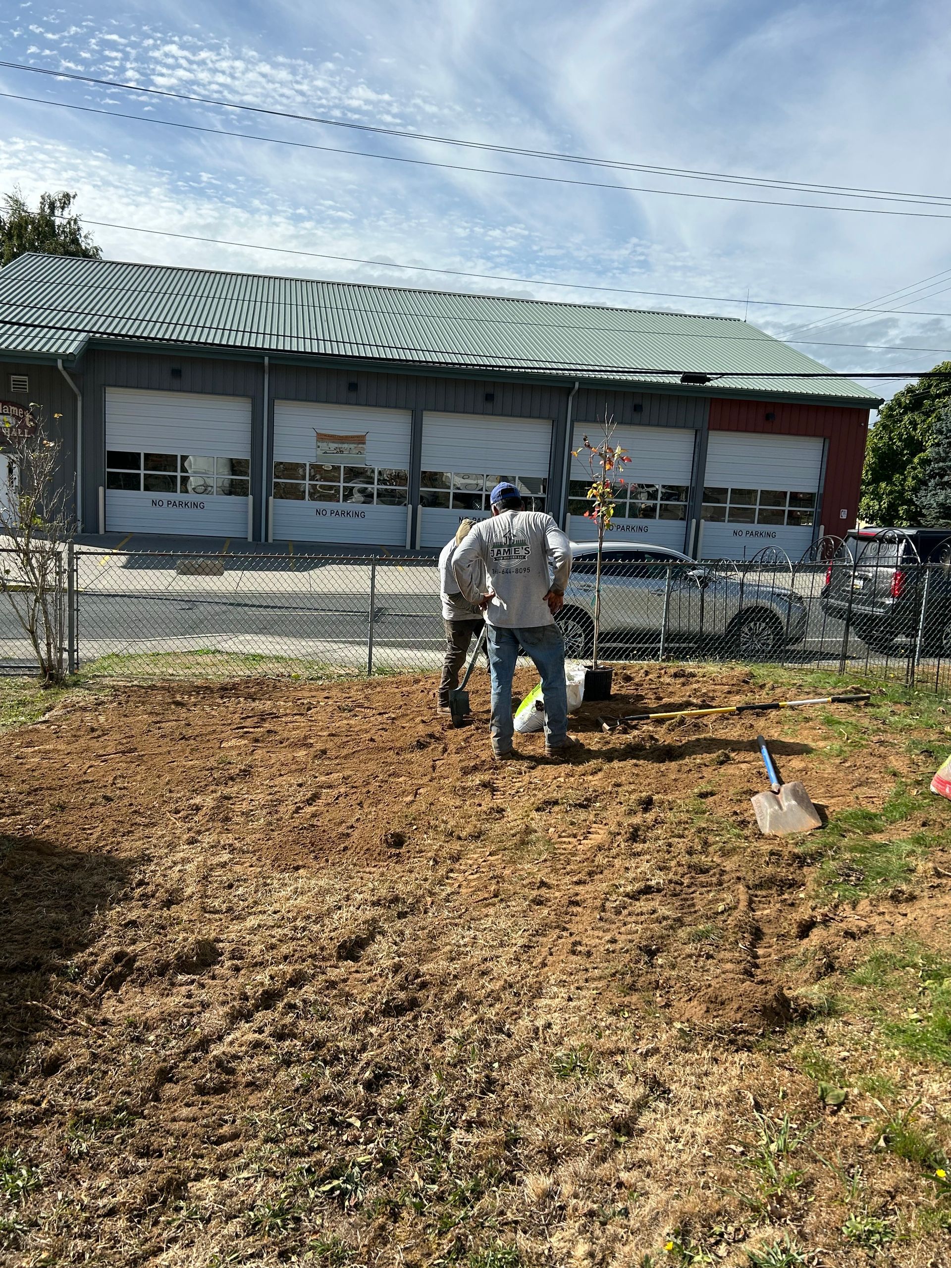 Two workers tending a muddy field beside a metal-roofed building under a cloudy sky.