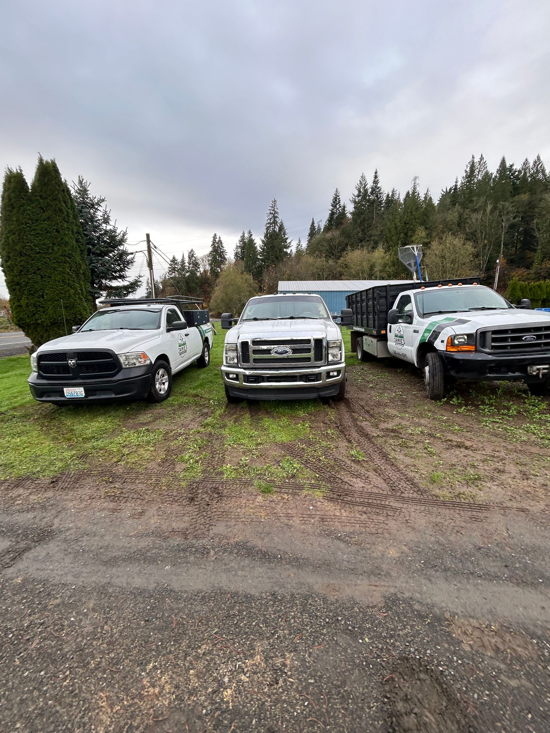 Three white pickup trucks parked on a muddy lot with trees and cloudy sky in the background