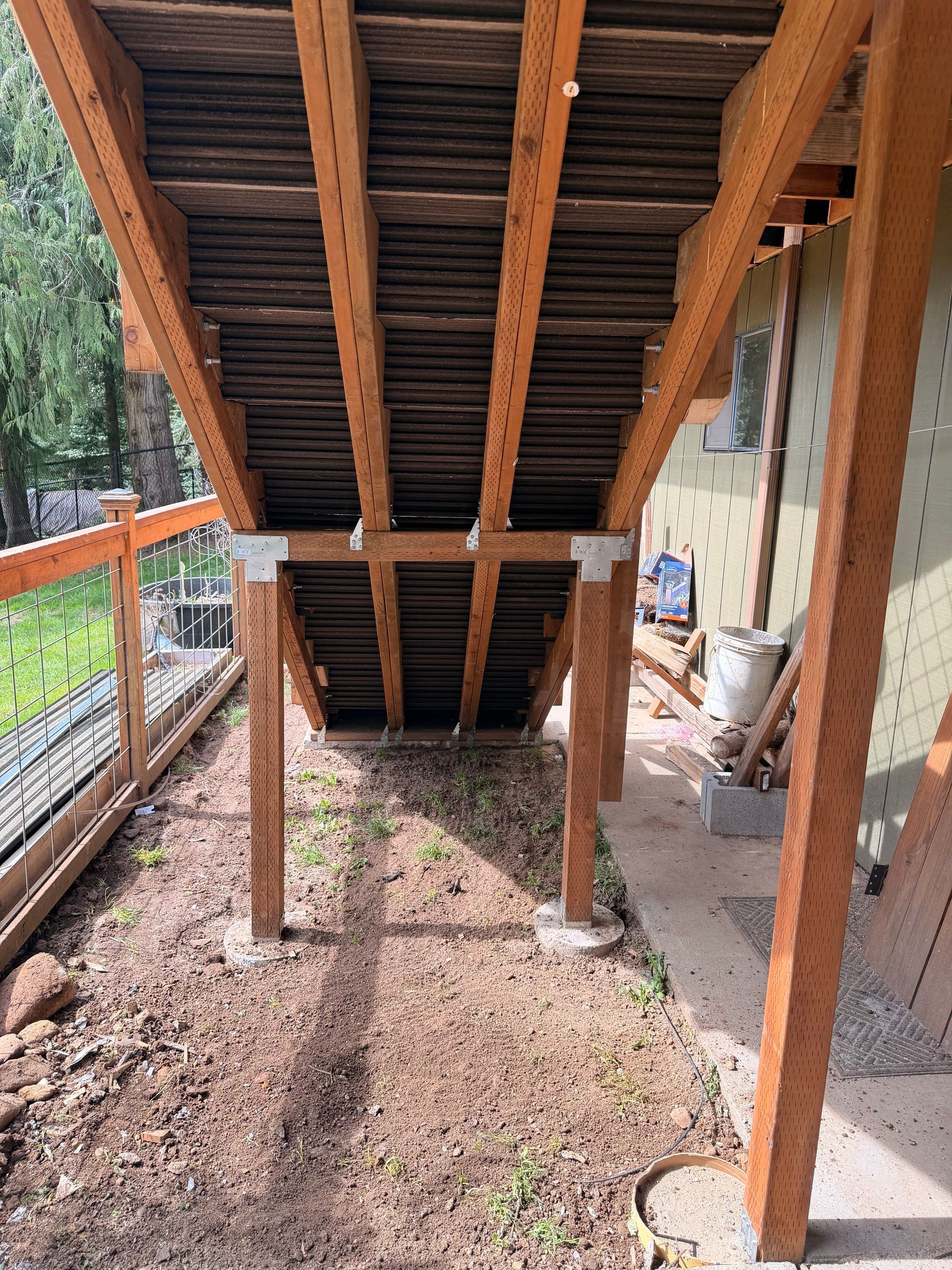 Wooden staircase and support posts on a deck, viewed from below, with dirt ground and railing beside it