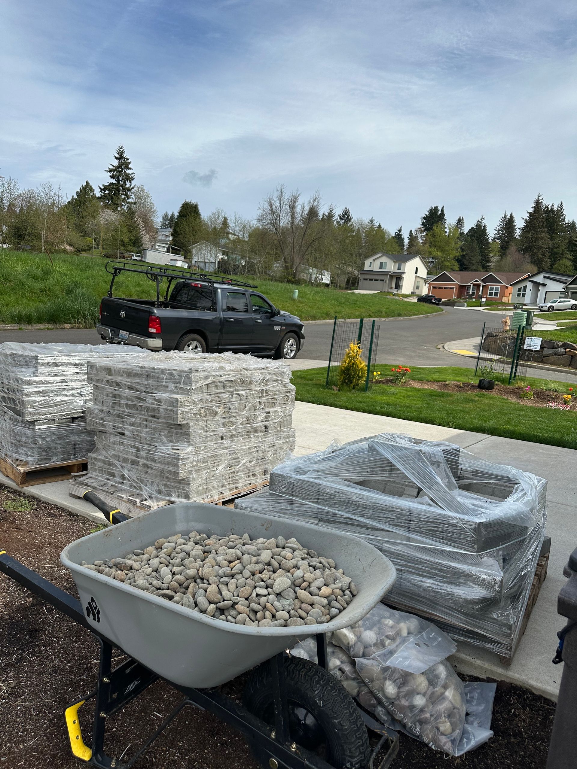 Wheelbarrow full of white stones beside stacked pallets in a yard, with a black pickup and road in the background