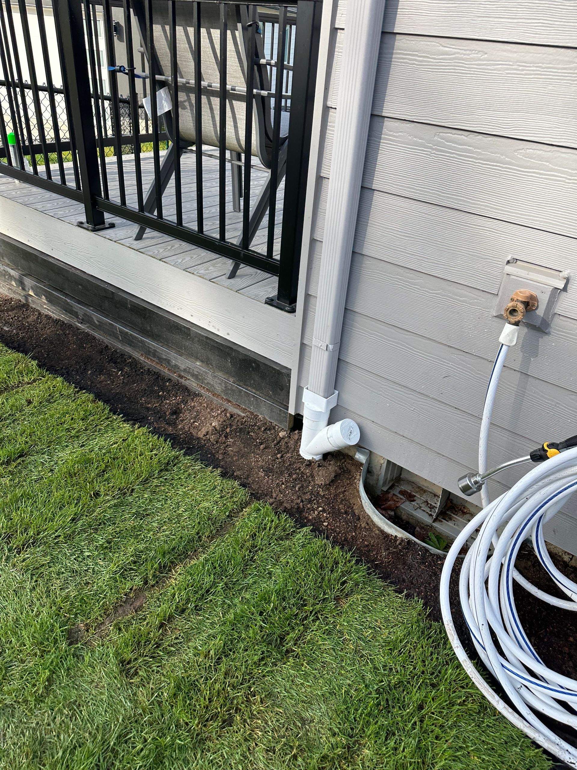 Exterior wall, fence, downspout, lawn edge, and coiled hose near a house corner.