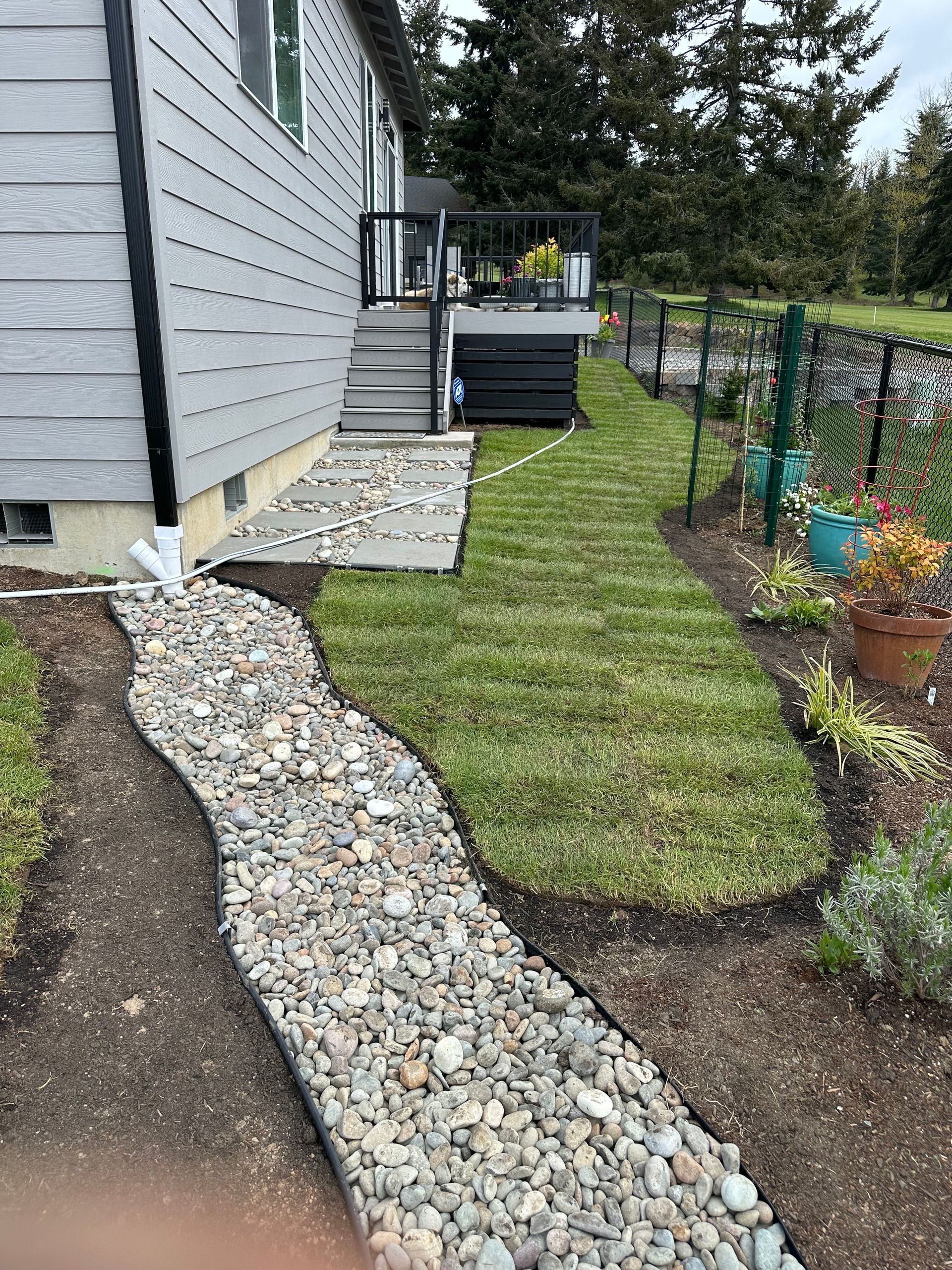 Narrow gravel garden path beside a gray house, bordered by mulch, lawn, and potted plants.