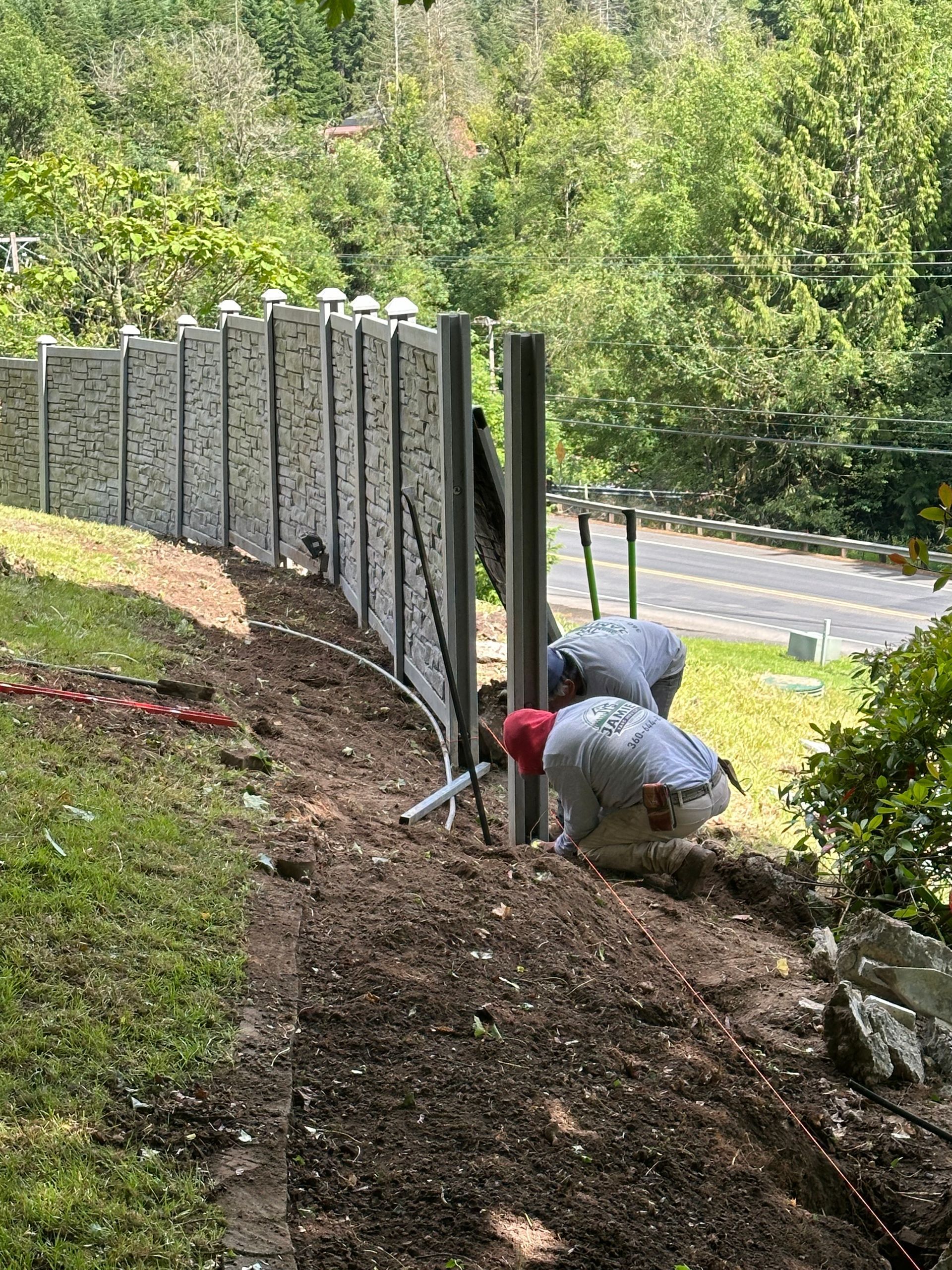 Worker in gray and red crouches by a fence, digging beside a road in a wooded area.