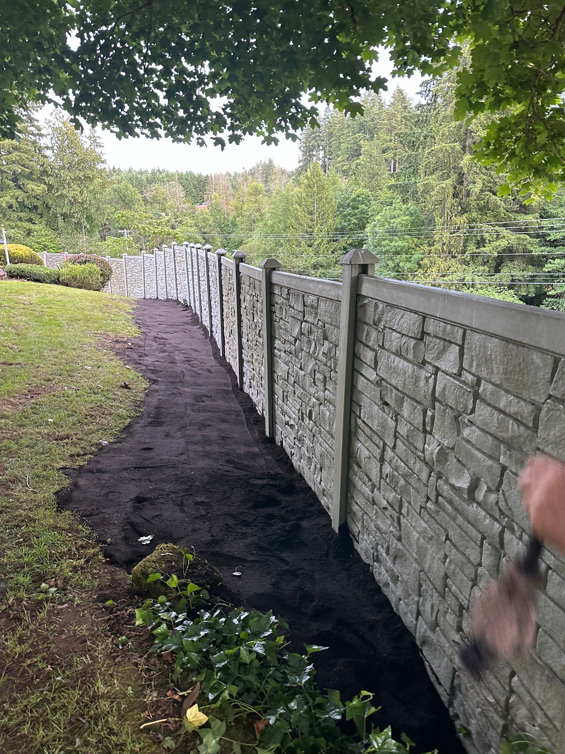 Dirt path beside a stone wall, with fresh black mulch along the edge under overhanging trees.