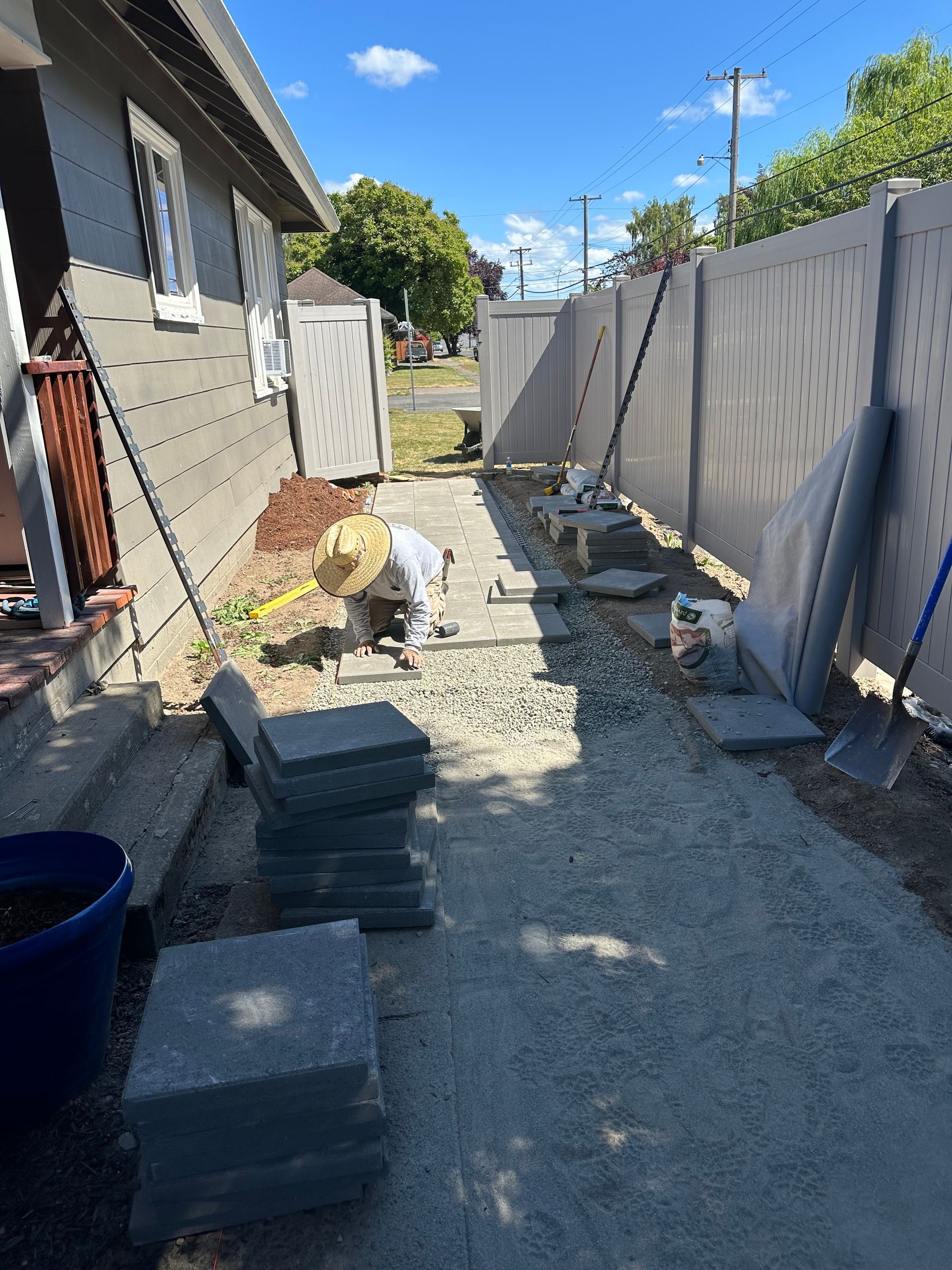 Narrow gravel walkway beside a house and fence, with stacked pavers and construction materials.