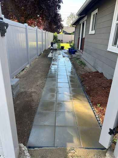 Wet concrete walkway beside a house and white fence, with yard tools and a blue bin at the far end