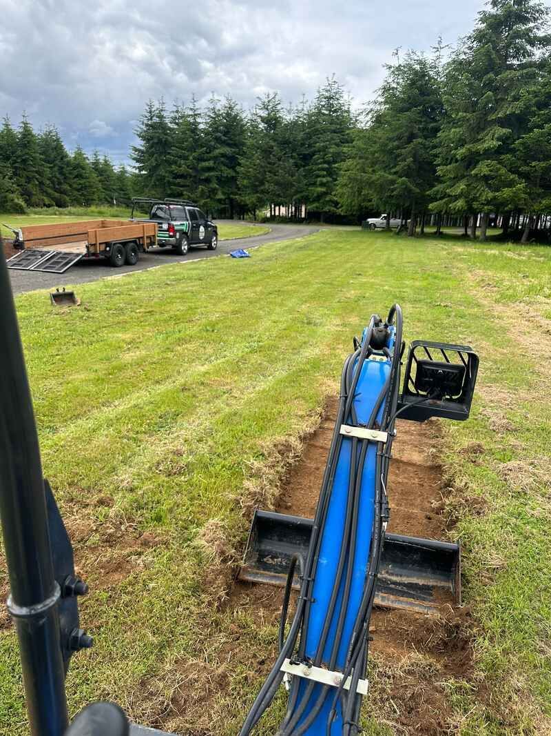 Blue post driver excavator attachment digging narrow trenches in a grassy field with trees in the background