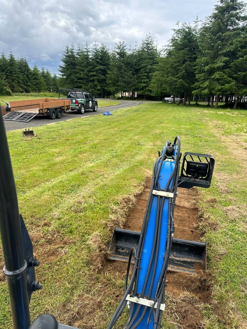 Blue excavator arm on a grassy field with trucks and trees in the background