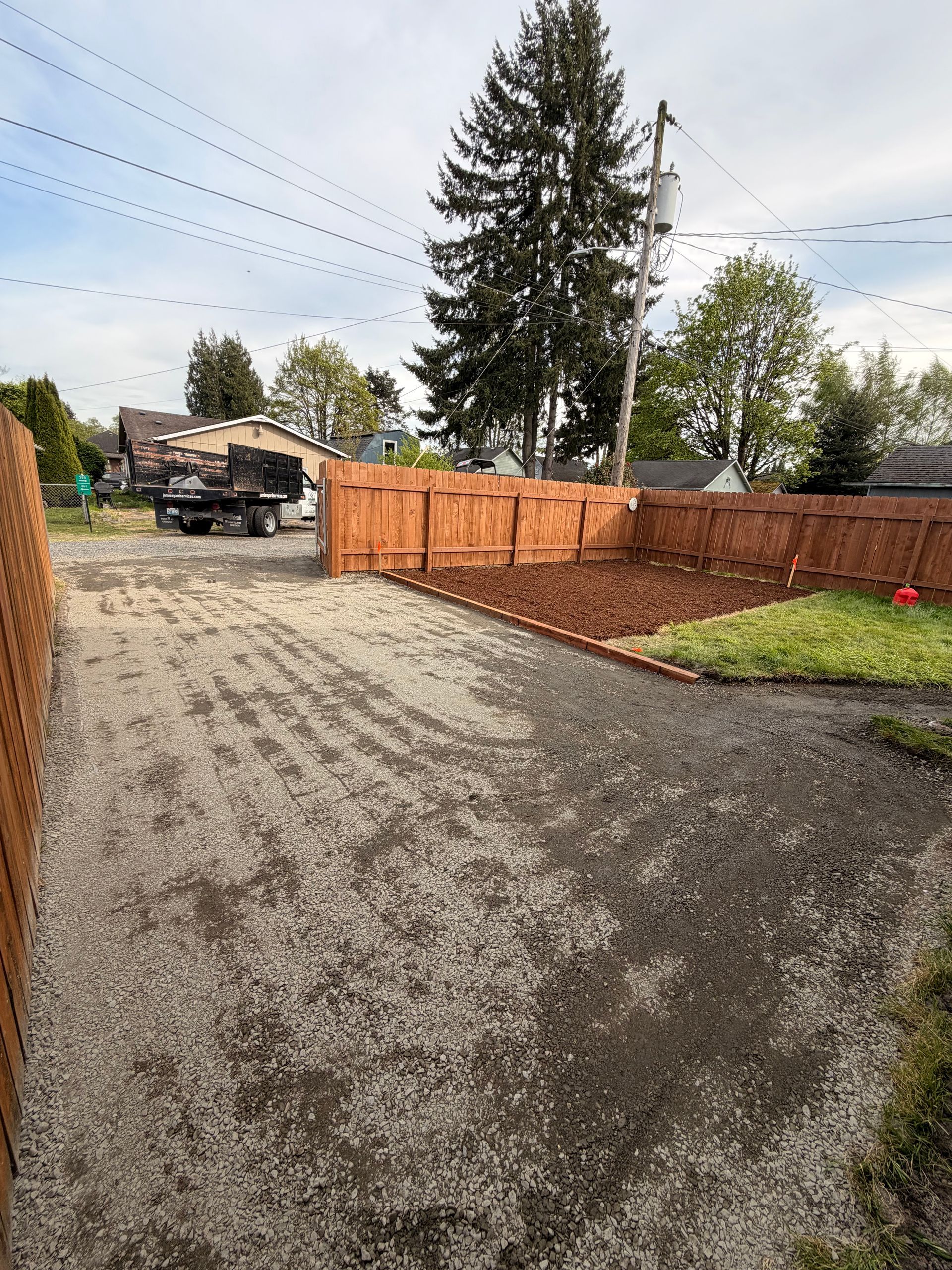 Gravel driveway beside a wooden fence and large evergreen trees under an overcast sky.