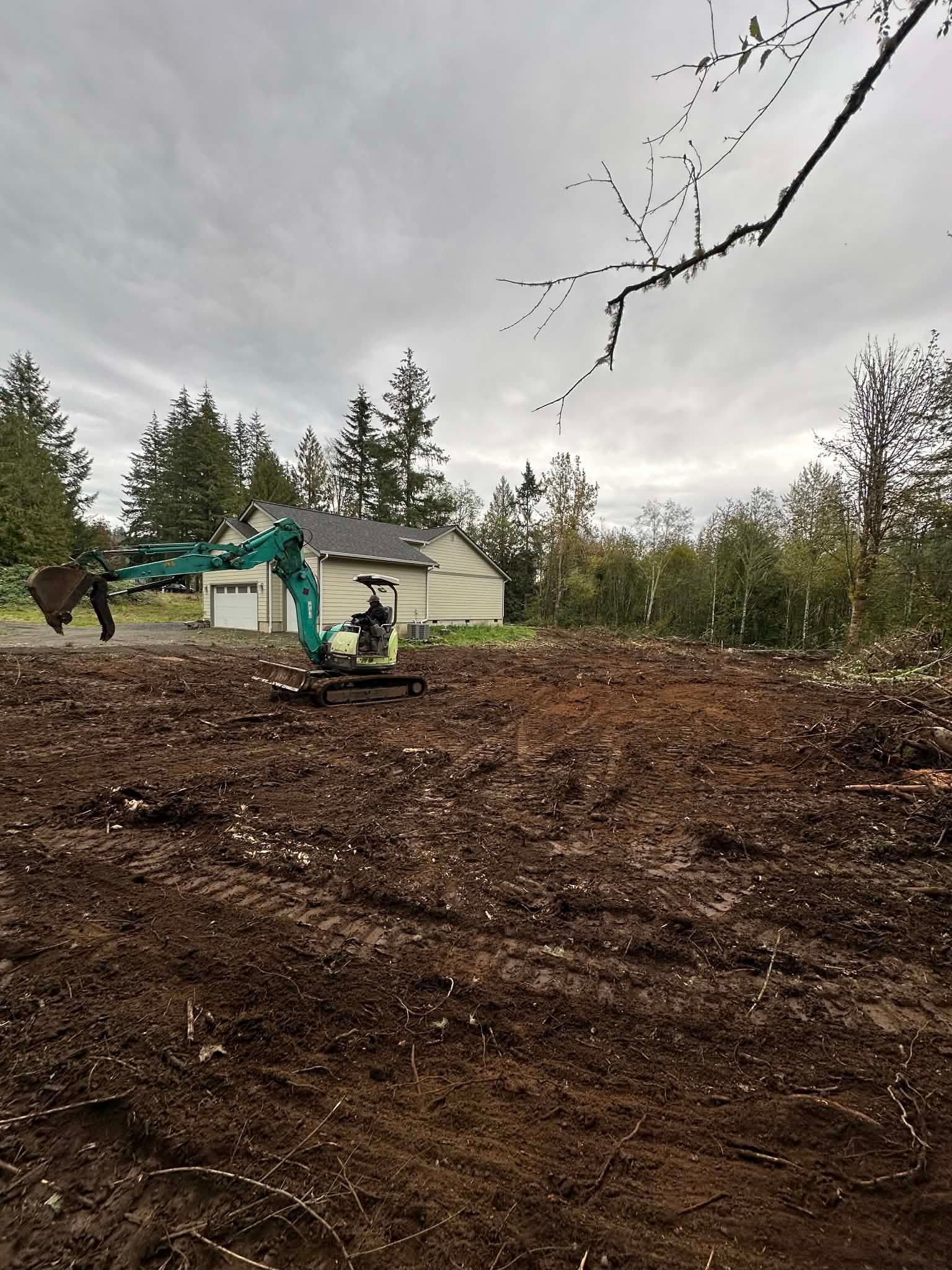Excavator clearing muddy dirt at a rural site with a small building and overcast sky