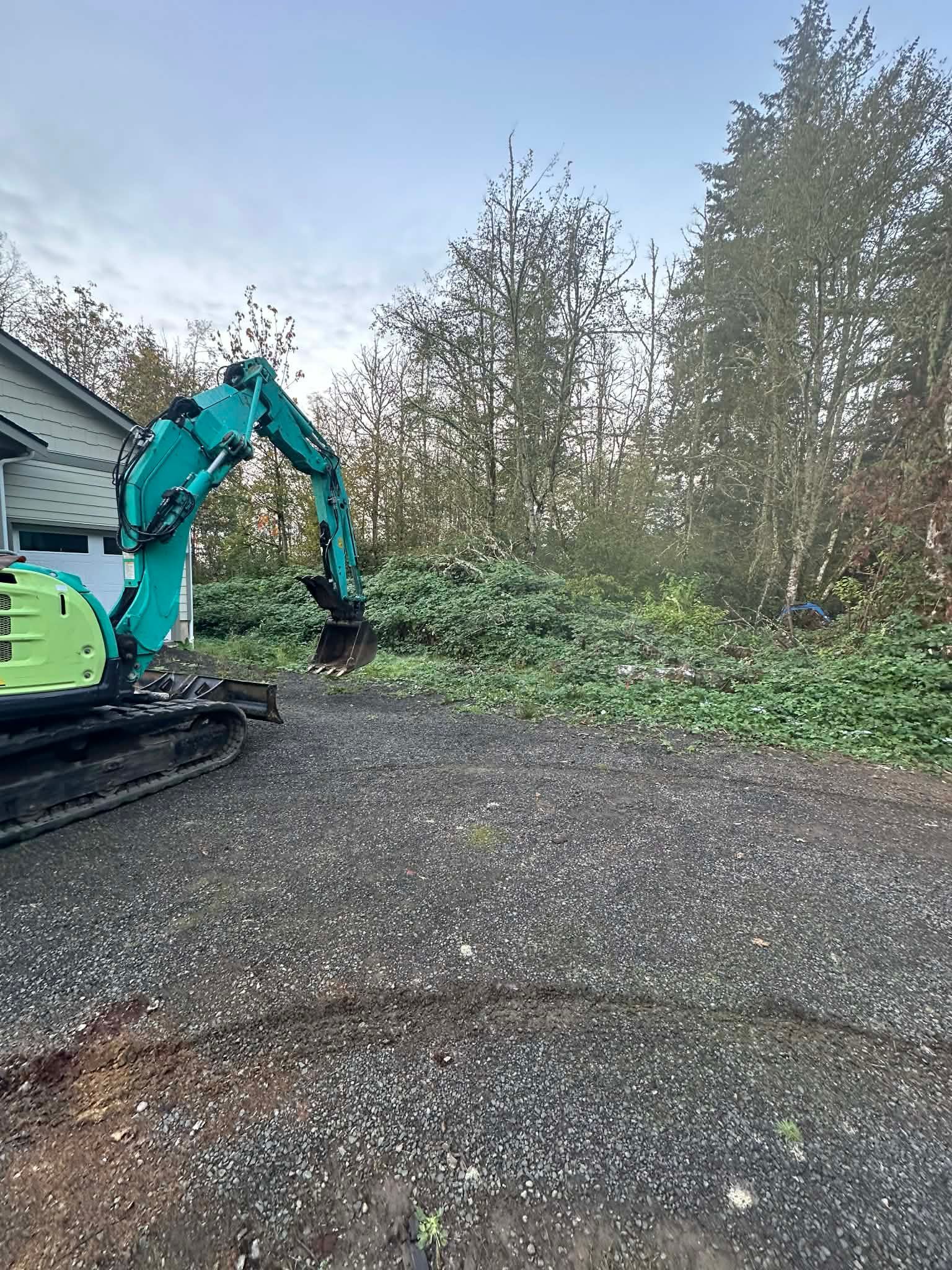 Turquoise excavator clearing brush beside a gravel lot and building