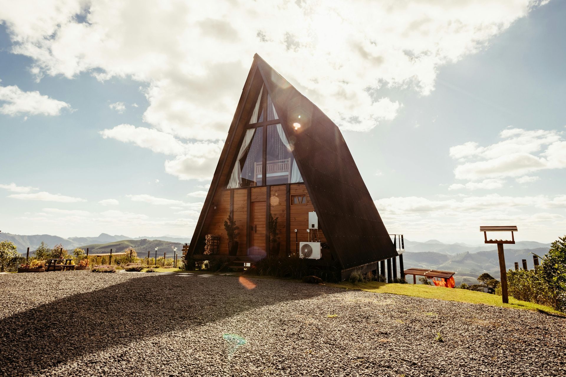 A modern A-frame cabin on a gravel hill under a bright, cloudy sky, with distant mountains.