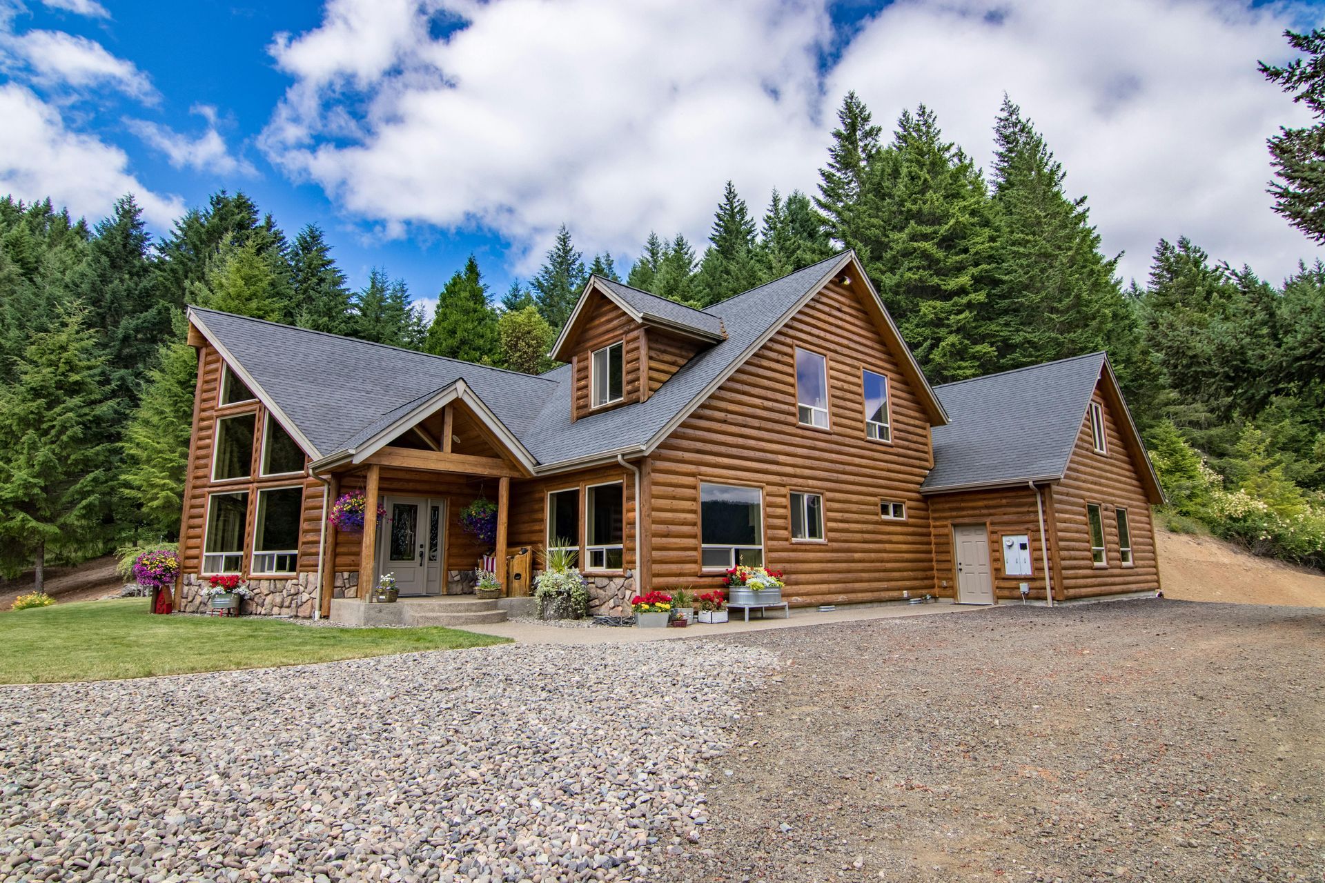 Rustic log cabin with stone accents and gravel driveway, surrounded by evergreen trees under a partly cloudy sky