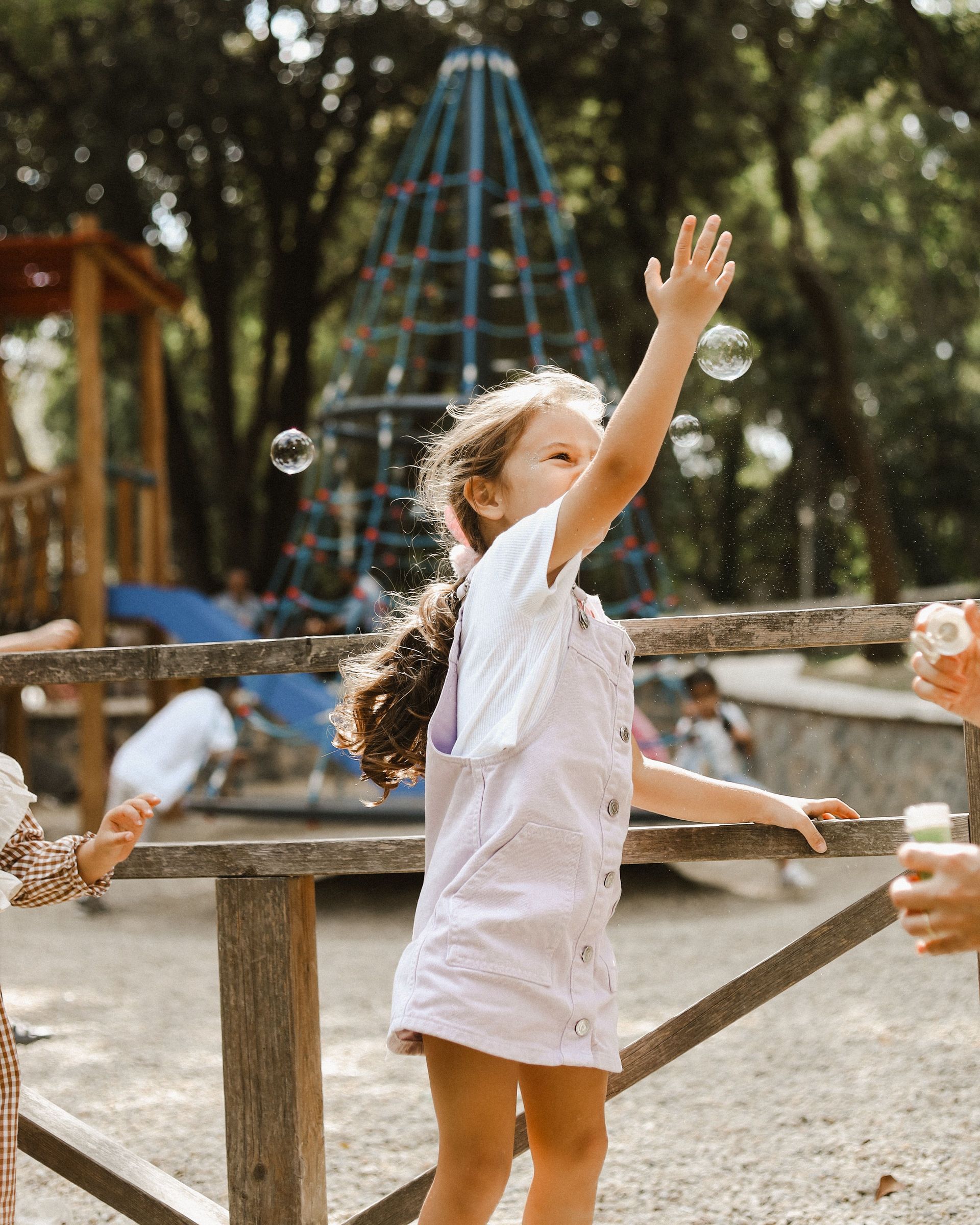 Child reaching up toward bubbles in a playground, with a climbing net in the background.