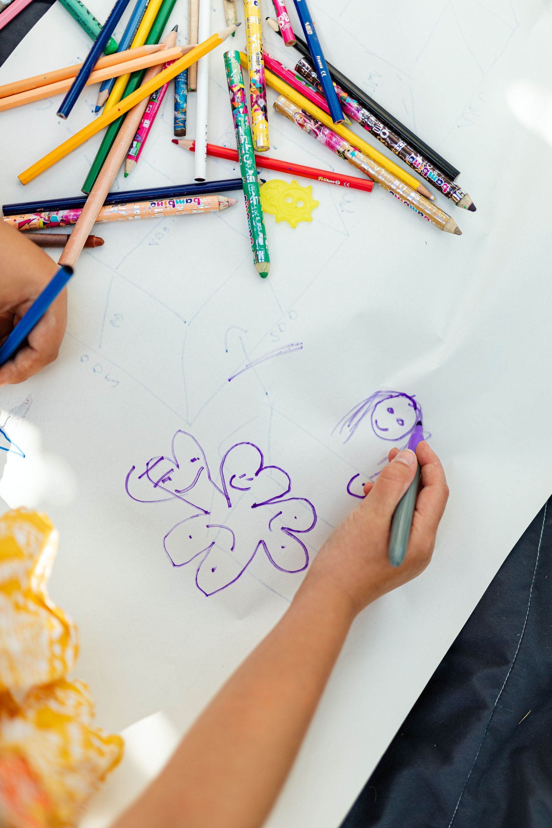 Child drawing flowers with crayons on a white table, with scattered colored pencils around.