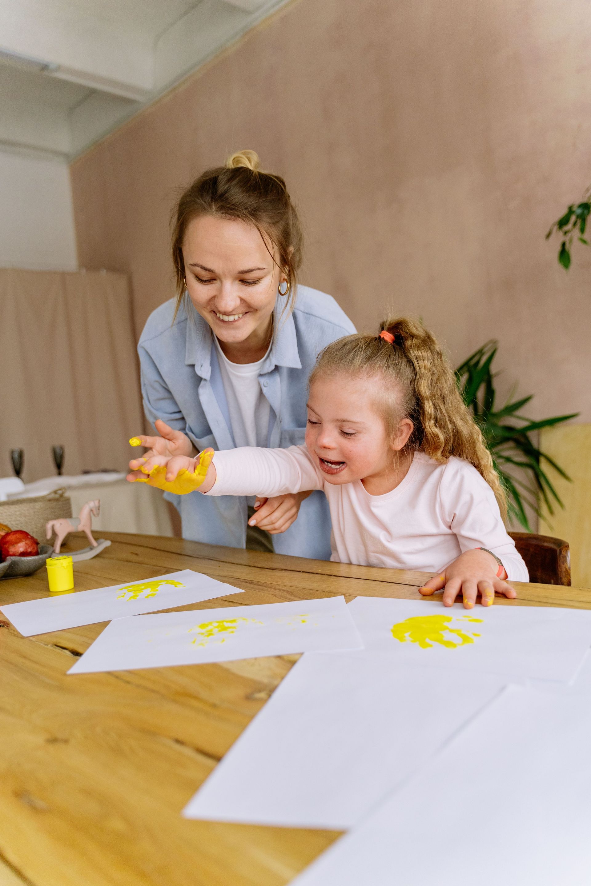 Woman and child painting at a table with paper and yellow paint, smiling together