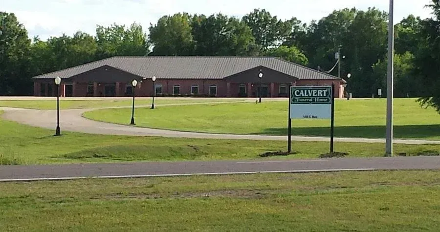 Single-story brick building behind a grassy field with lampposts and a green sign near the road.