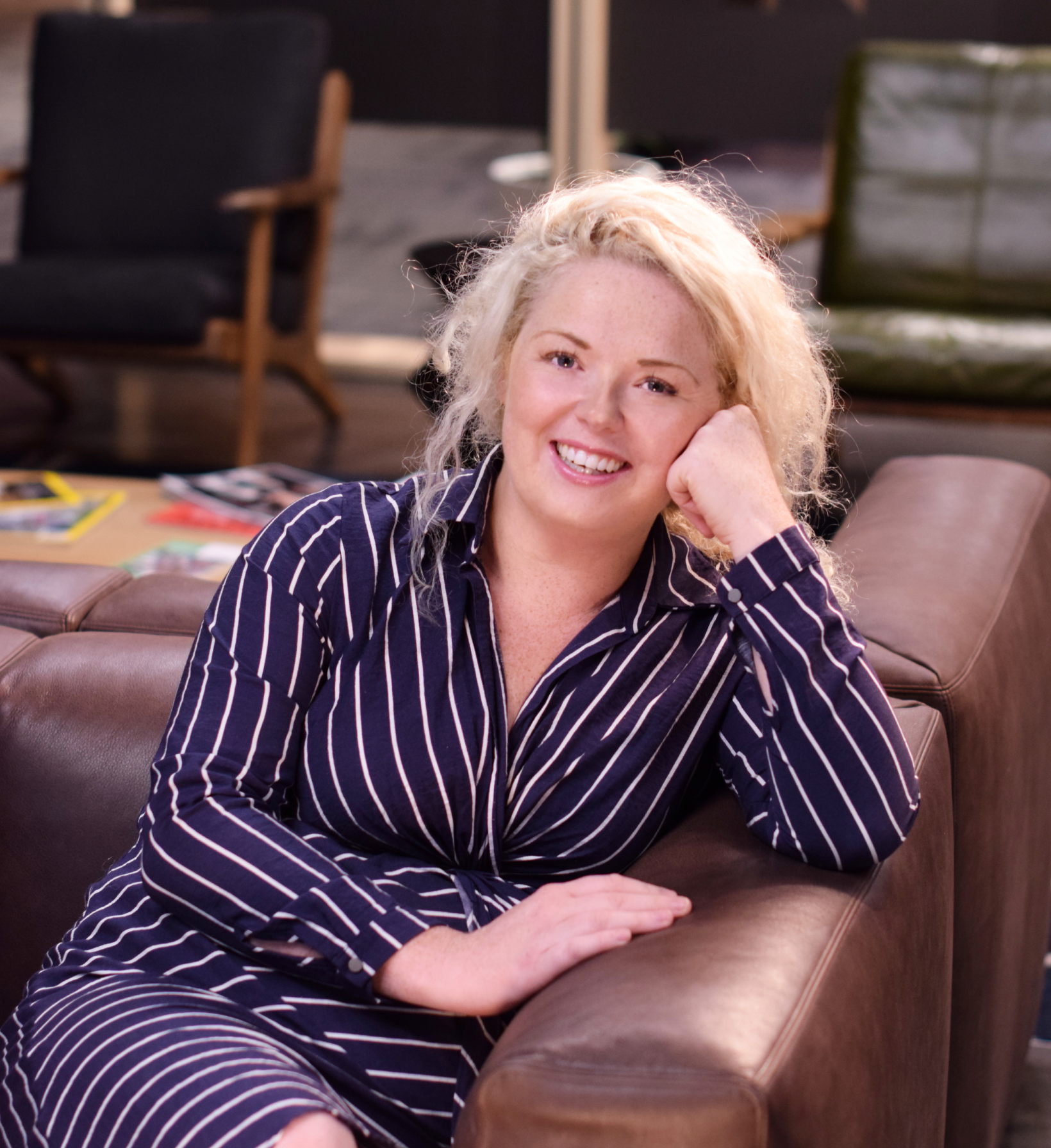 Smiling blonde woman in a navy striped shirt reclining on a brown leather sofa