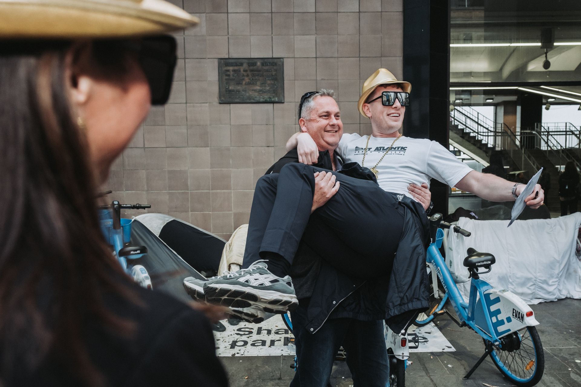 Two smiling men on a small bike, one carrying the other, outdoors near a building