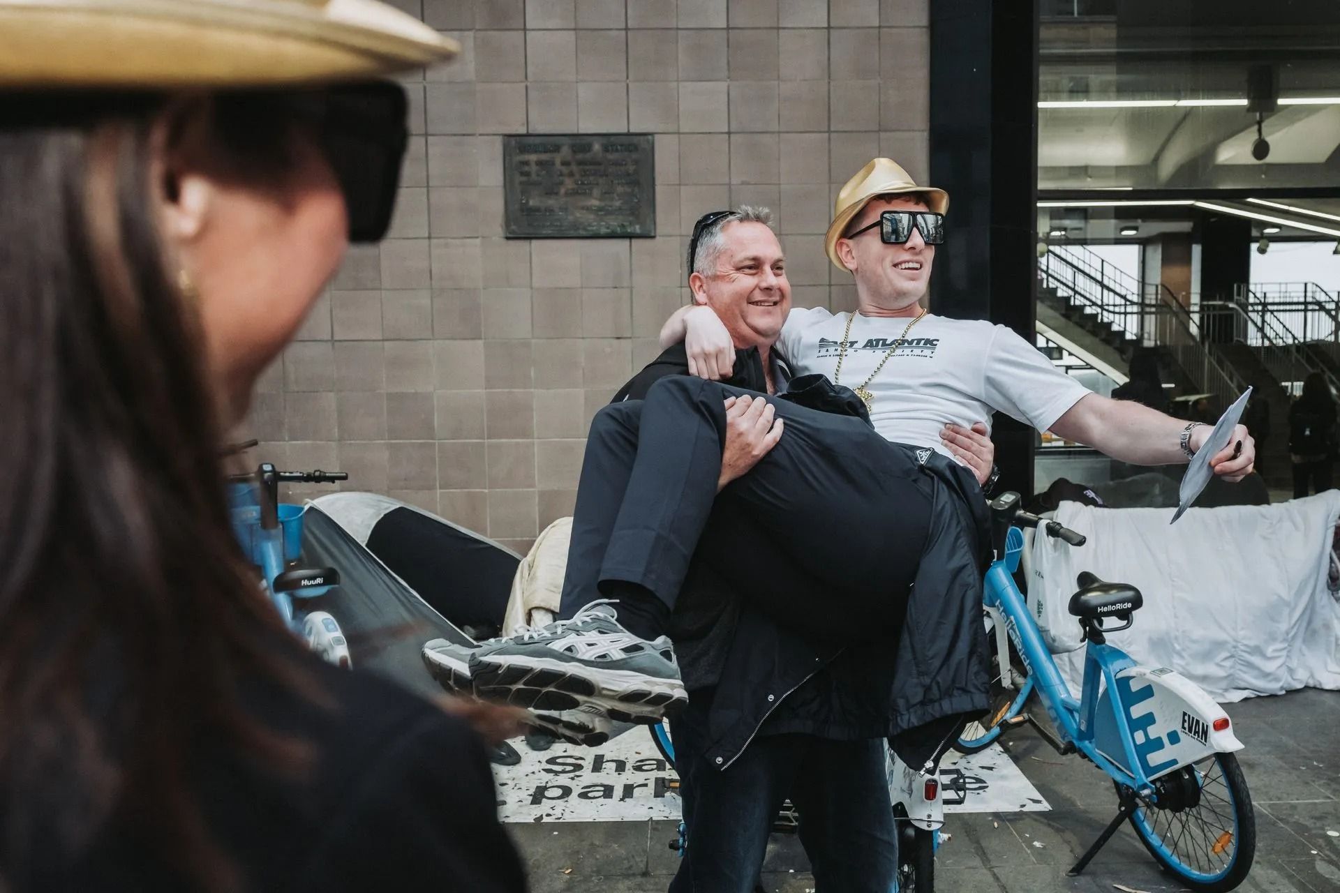 Two smiling people on a small blue bike, one carrying the other, in front of a building.
