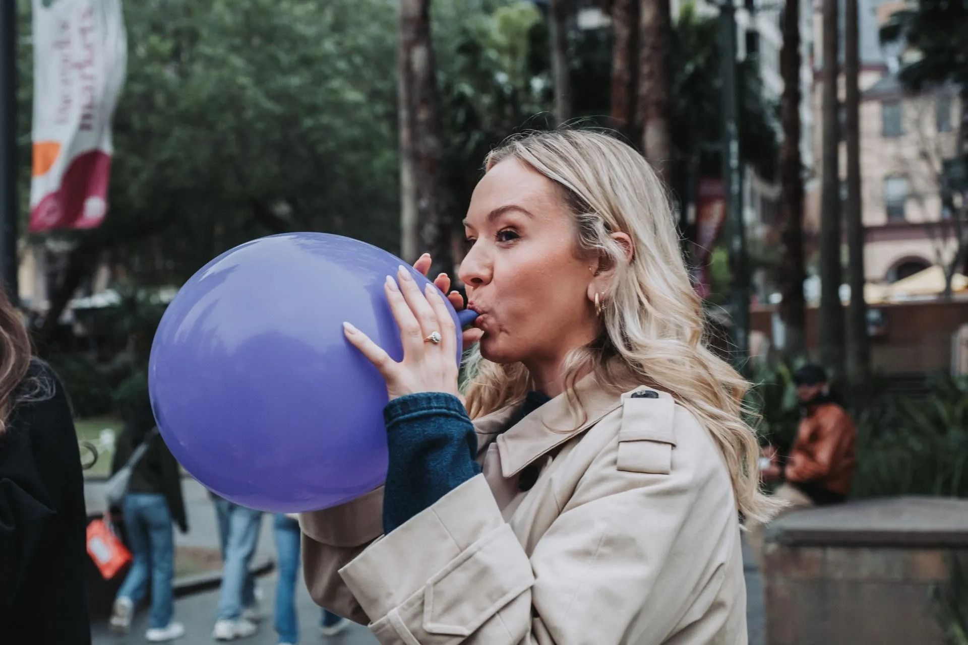 Woman in a beige coat blowing into a purple balloon outdoors in a park-like setting