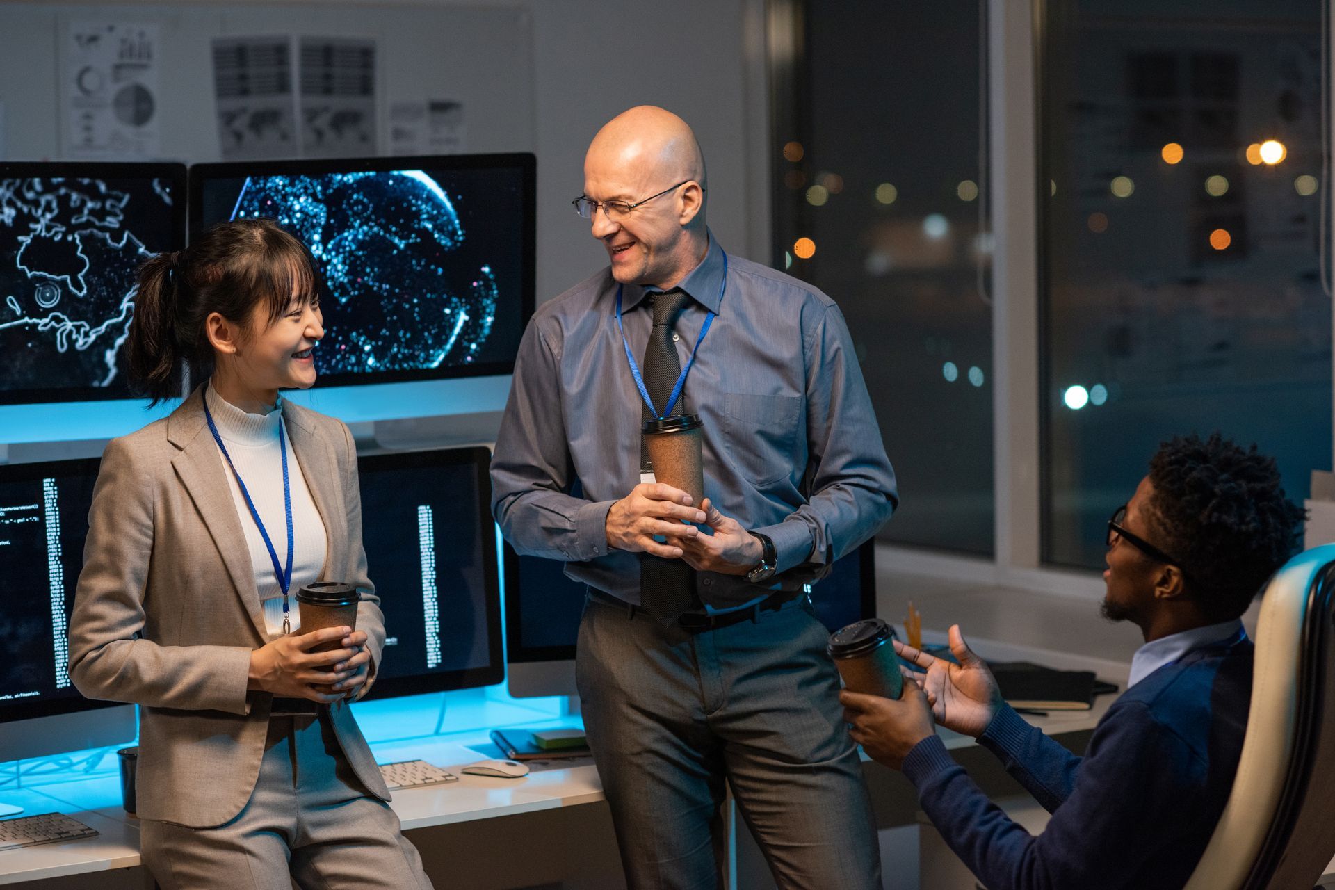 Three coworkers talking in a dim office near glowing monitors and a world map display.