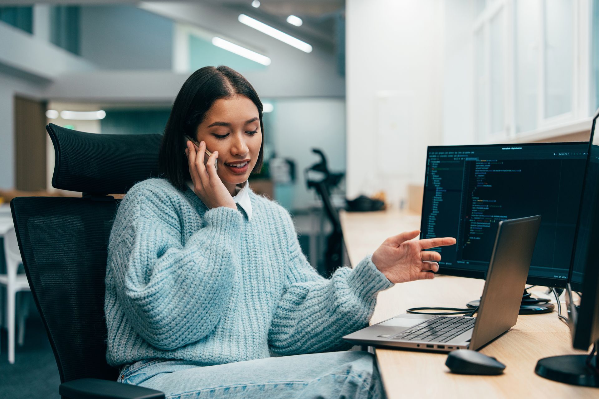 Woman in a light blue sweater talks on the phone while pointing at a laptop in a modern office.