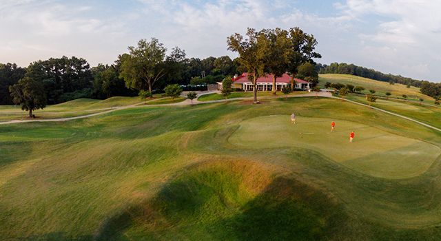Rolling green golf course with a clubhouse in the distance under a blue sky.