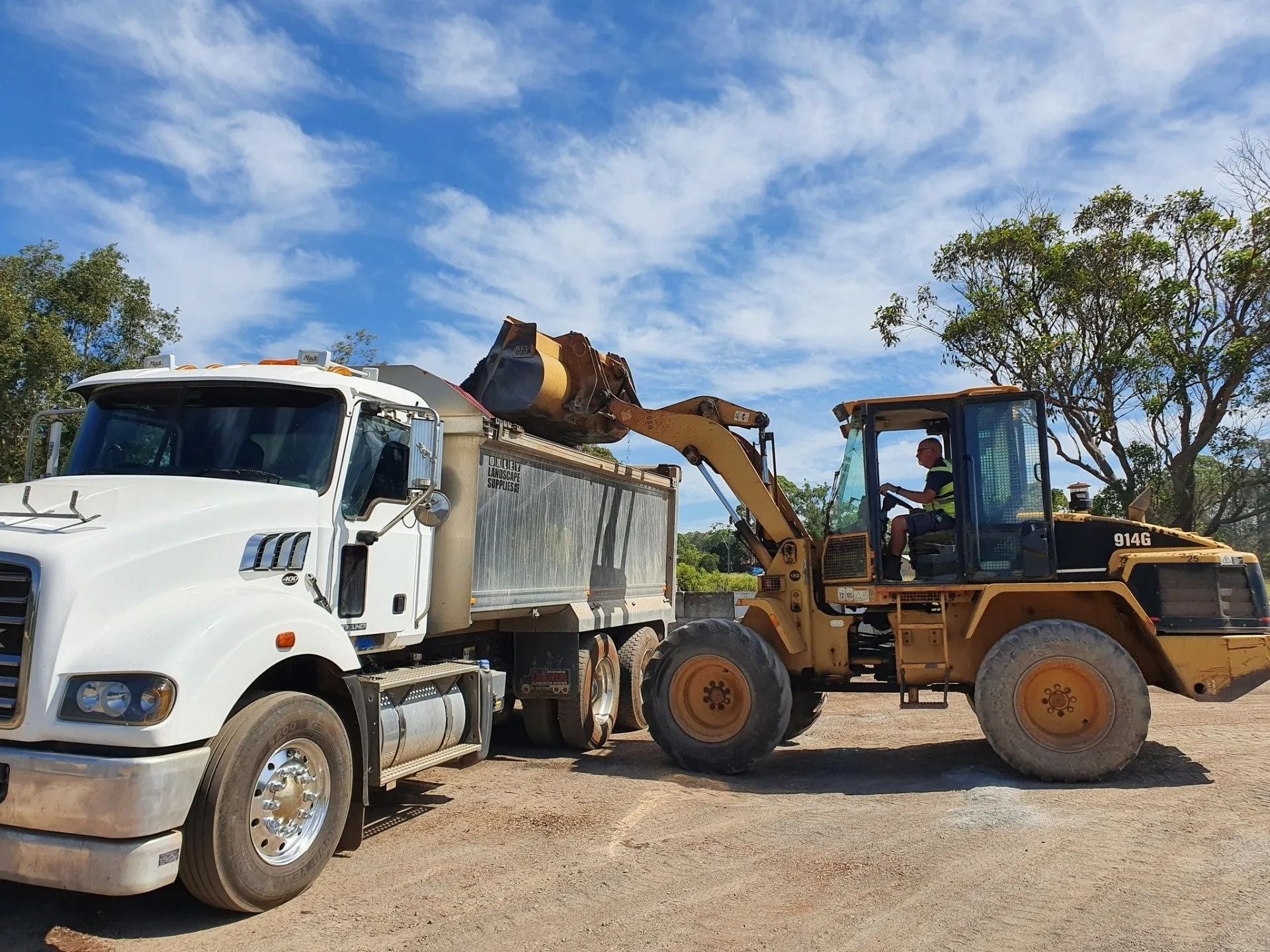 A Yellow Loader Dumping Material Into a White Dump Truck, on A Dirt Road — Port Landscape Supplies in Port Macquarie, NSW