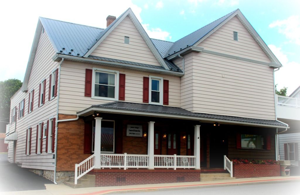 Two-story building with a porch and brick base, tan siding, burgundy shutters, and a gray metal roof.