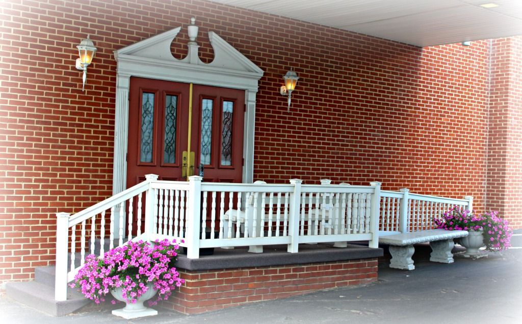 Brick building entrance with white railing, steps, and a red door; purple flowers in pots.