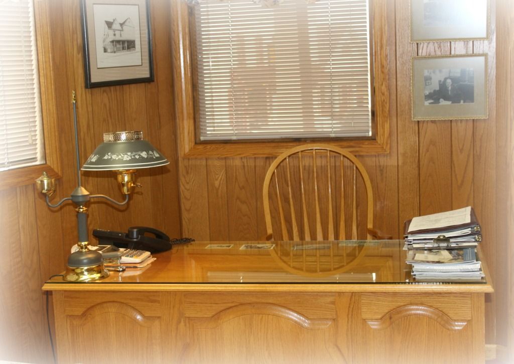Wooden desk in an office with a lamp, chair, and window. Photos hang on the wood-paneled walls.