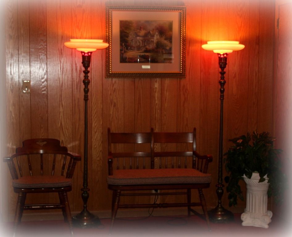Two ornate floor lamps flank a bench and chair against a wood-paneled wall with a framed picture.