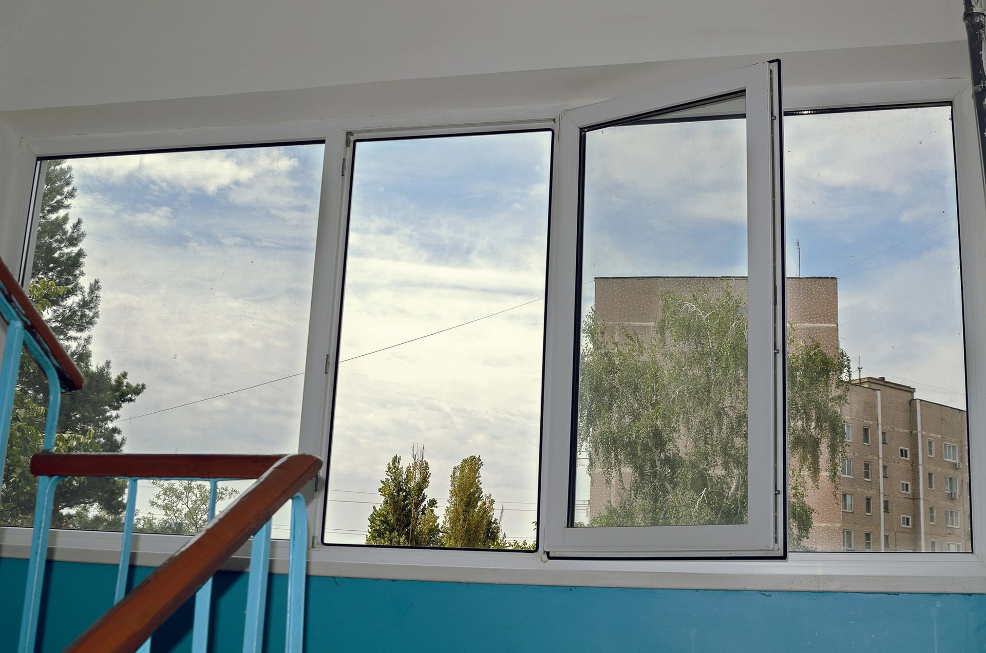 Finestra aperta con quattro vetri e una parete blu, che si affaccia su alberi e palazzi.