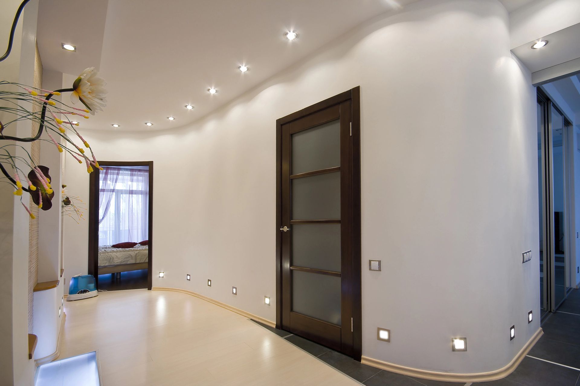Luminoso corridoio moderno con pareti bianche, faretti a incasso sul soffitto, porta scura e pavimento lucido.